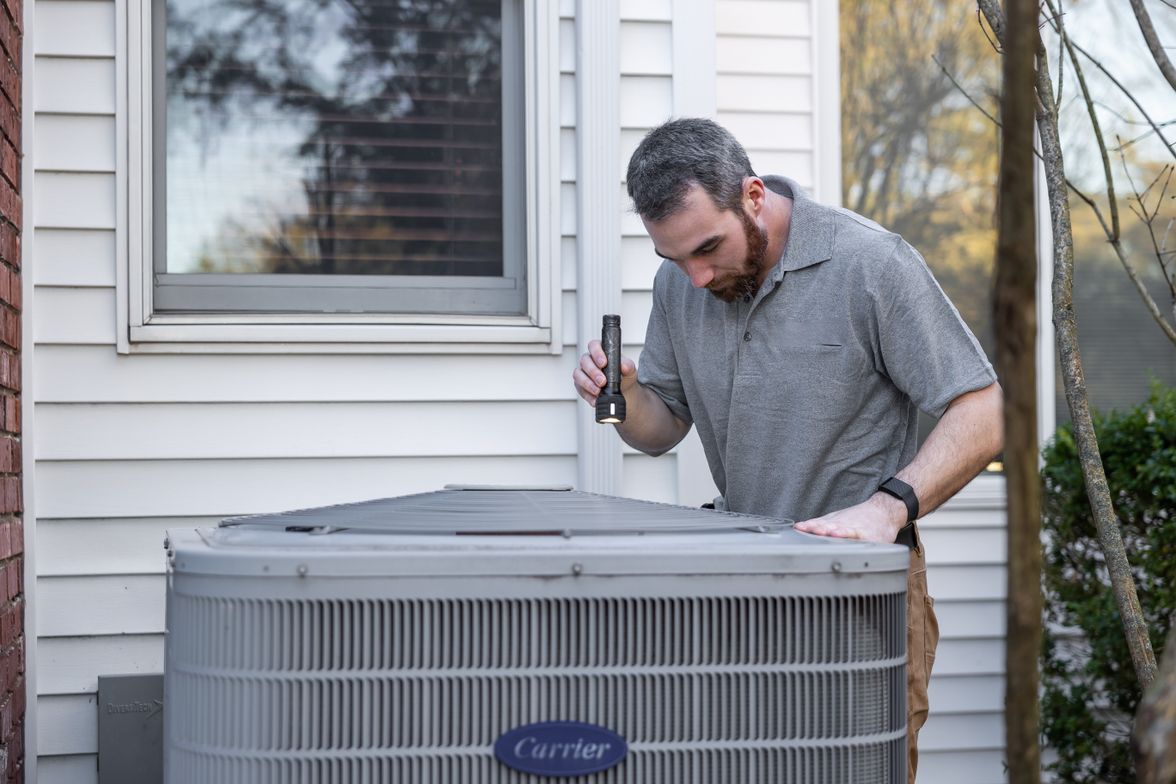 Technician with a flashlight inspecting a residential air conditioning condenser beside a house with white siding.
